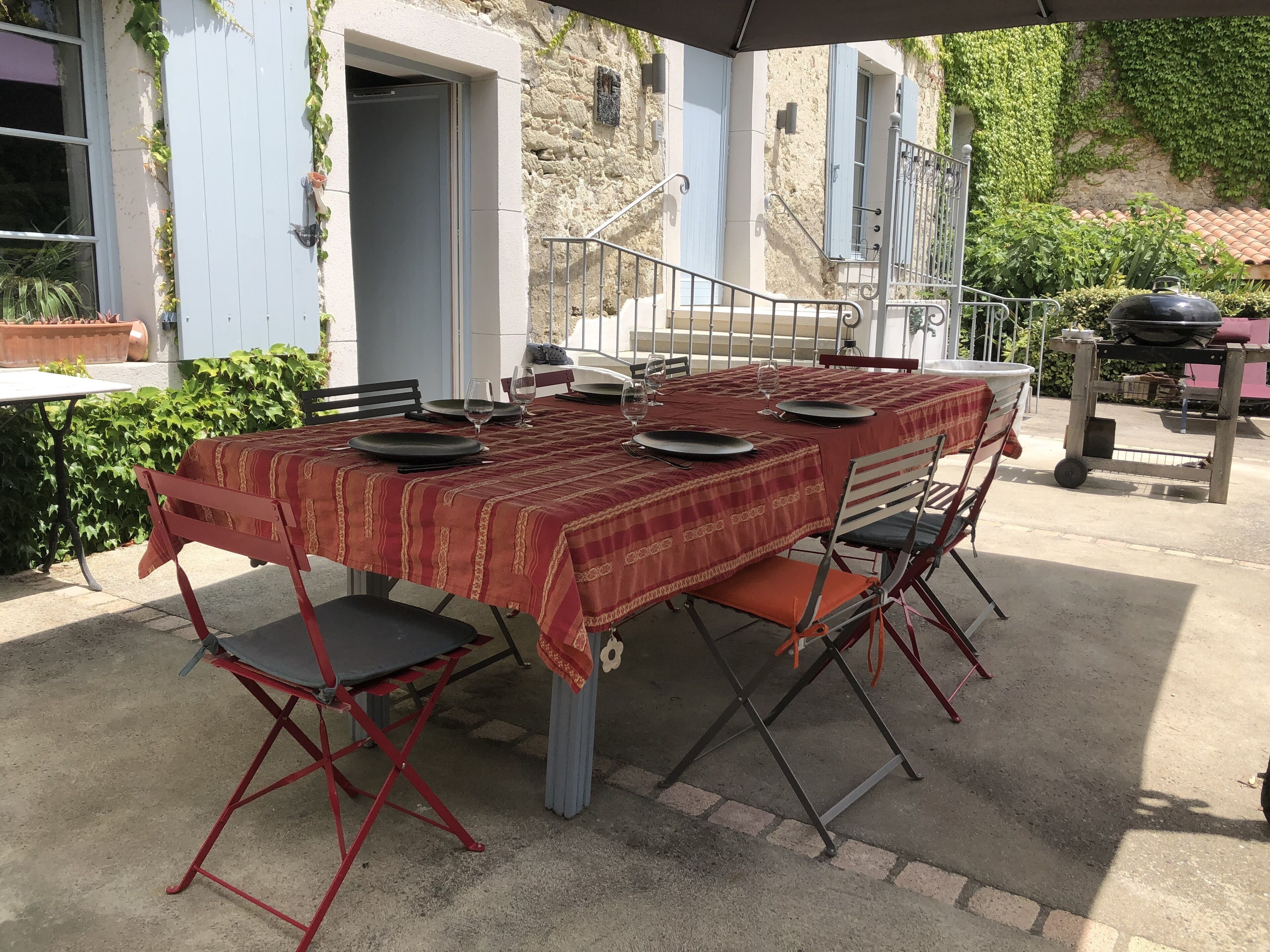 Un área de comedor al aire libre con una mesa preparada para cuatro personas, rodeada de sillas y una parrilla de barbacoa al fondo.