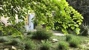A garden with a stone wall, various plants, and a building in the background. - Le Grenier Occitan (Gramazie)