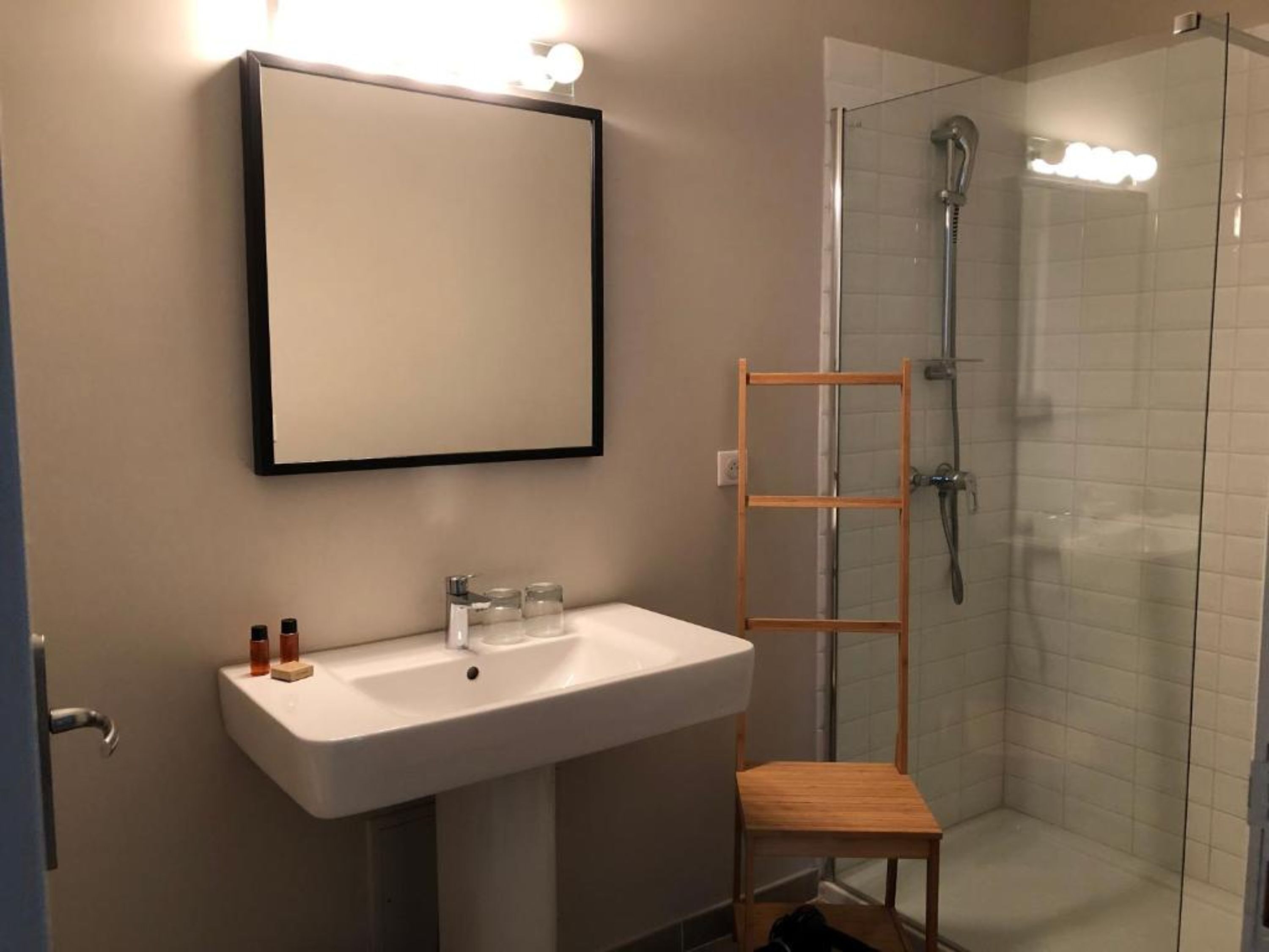 A bathroom with a white sink, a wooden shelf, a shower, and a mirror.