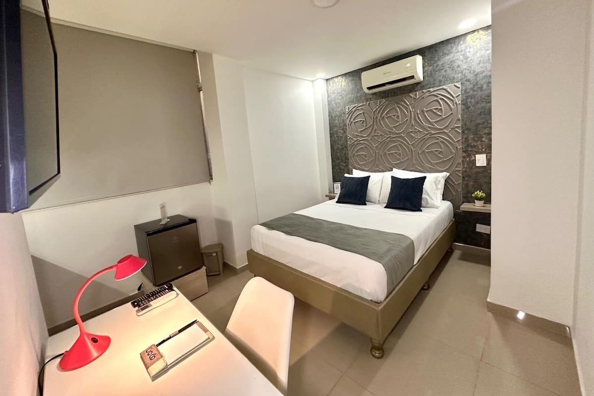 A hotel room with a bed, bedside tables, a desk with a lamp, and a wall-mounted air conditioner.