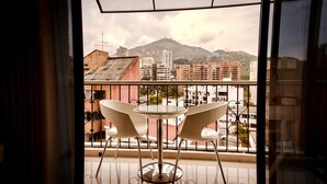 Un balcón con dos sillas blancas y una mesa redonda pequeña, con vista a un paisaje urbano con edificios y una montaña al fondo.