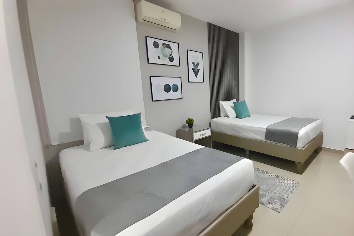 A hotel room with two beds, a small bedside table with a lamp, and a wall-mounted air conditioner.