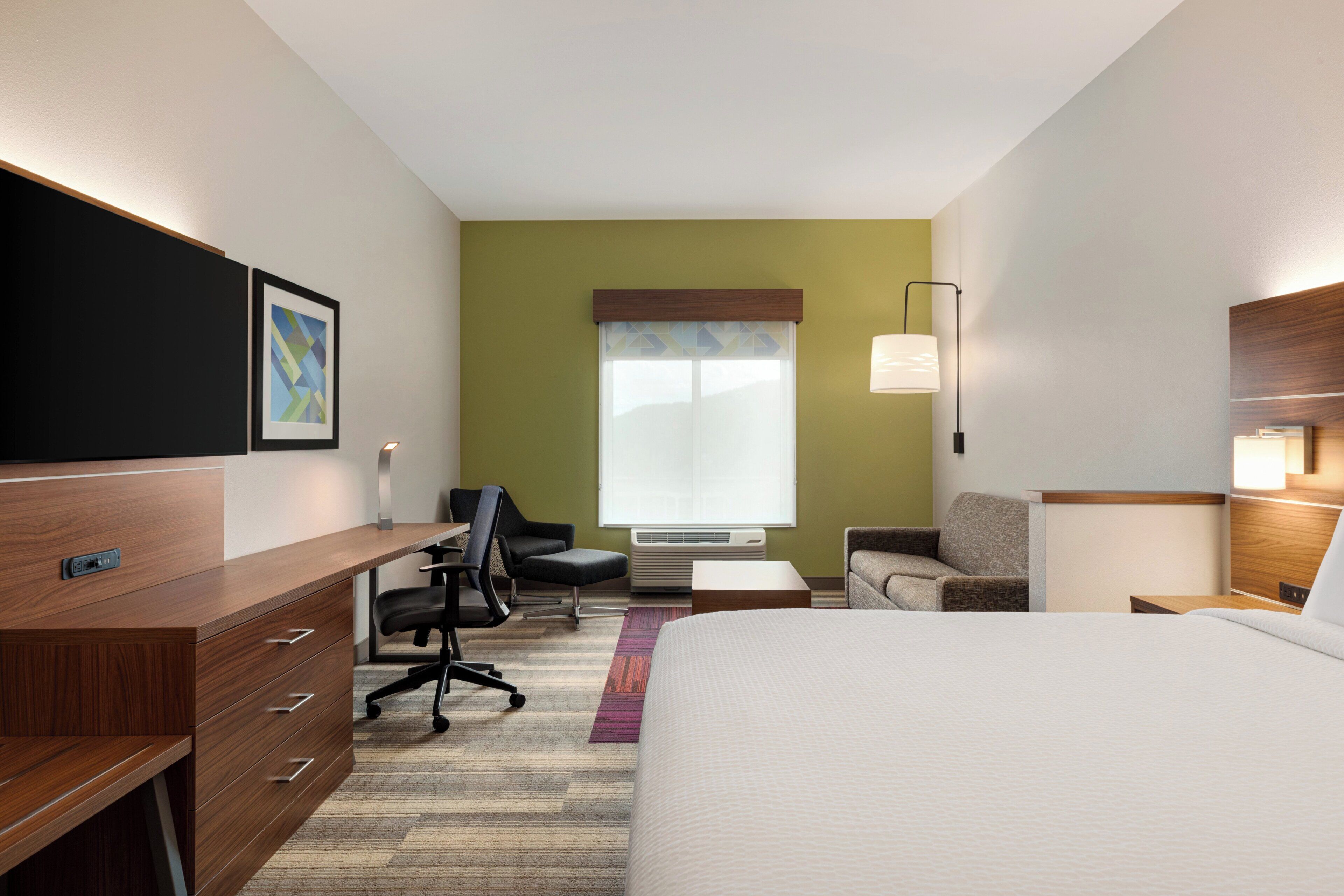 A hotel room with a bed, desk, chairs, and a window with blinds.