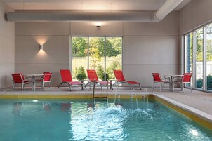 Una piscina interior con agua cristalina, rodeada de sillas y mesas rojas, ventanas amplias y vistas a zonas verdes al exterior.