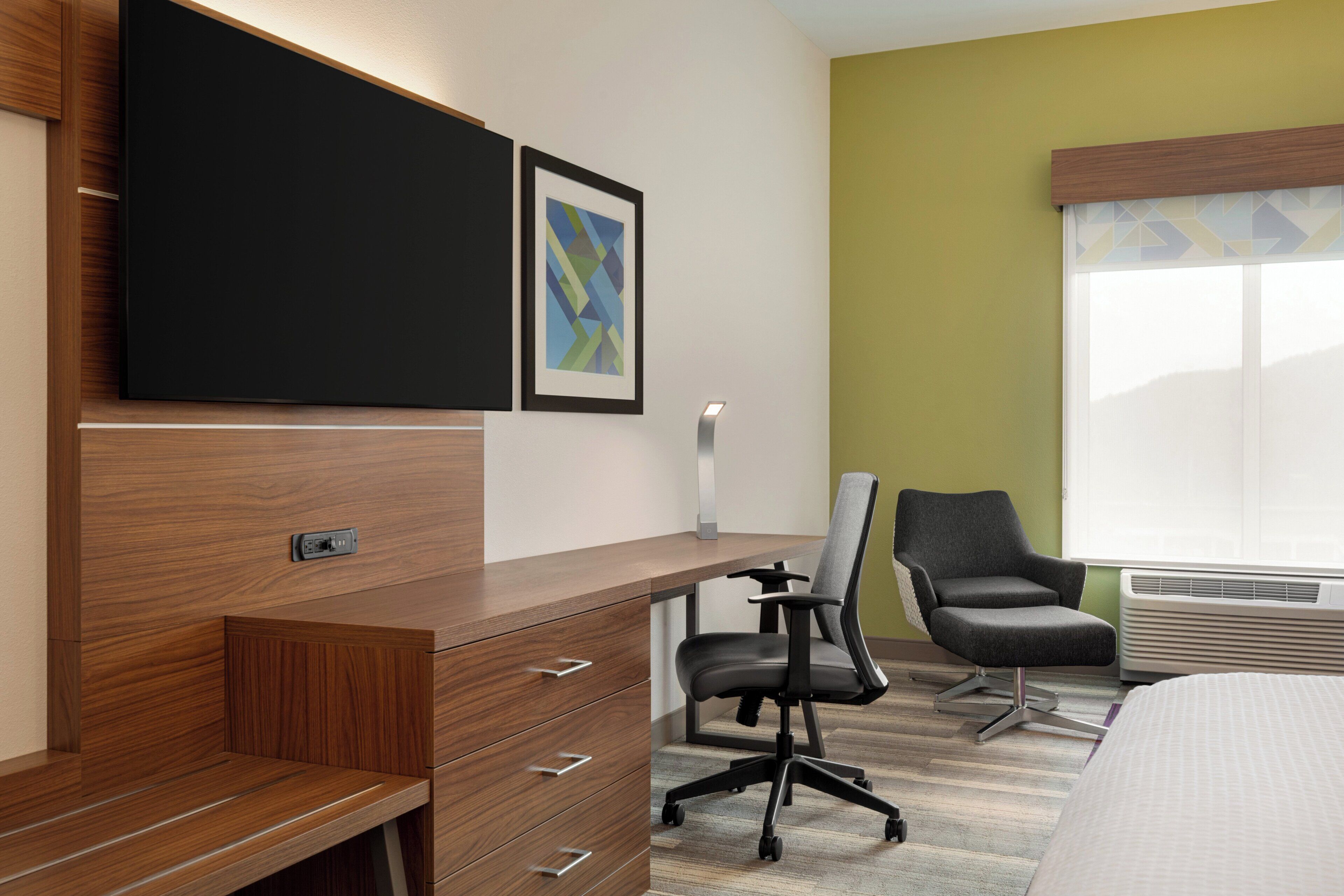 A hotel room with a wooden desk, a black office chair, a bed, a wall-mounted TV, and a framed abstract art piece.