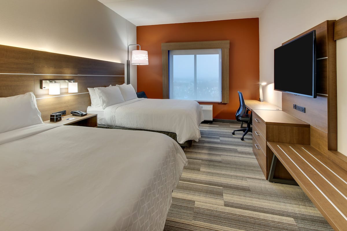 A hotel room with two beds, a desk, a chair, a flat-screen TV, and a window with a city view.