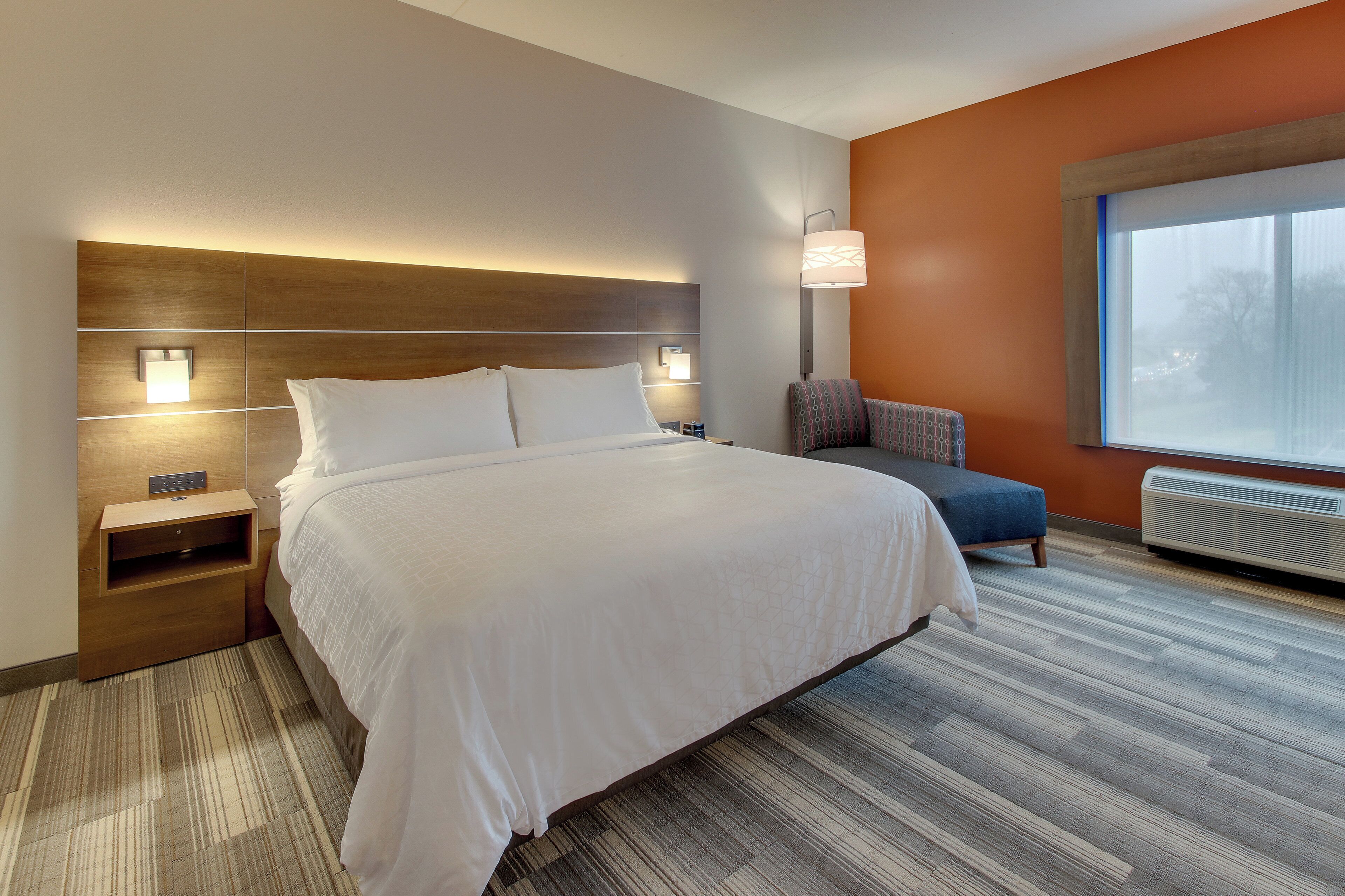 A hotel room with a large bed, bedside tables, a chair, a window, and a wall-mounted lamp.