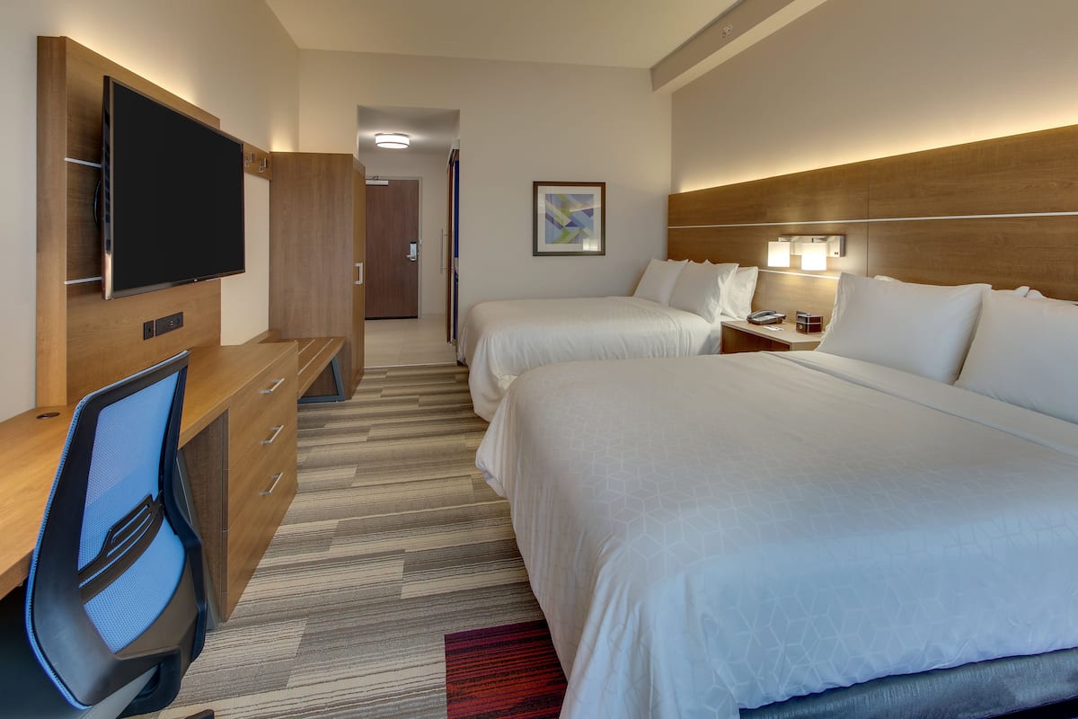 A hotel room with a large flat-screen TV, two beds, a desk with a chair, a nightstand, and a wall-mounted light fixture.