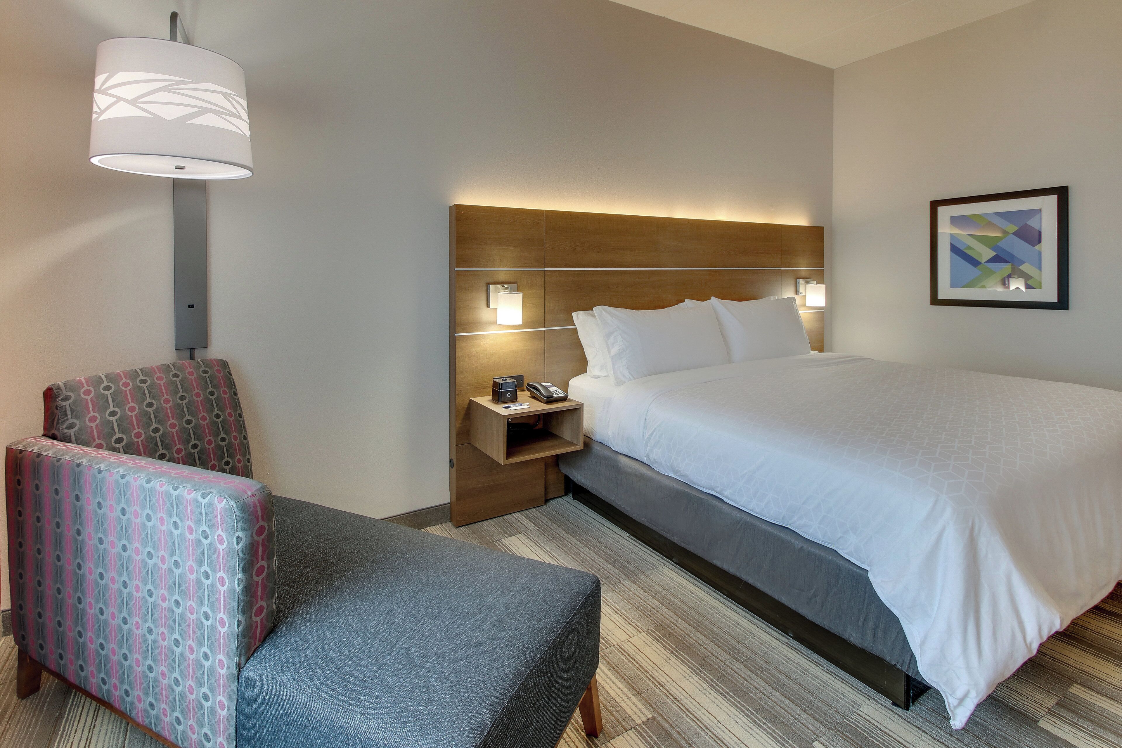A hotel room with a bed, a chair, a nightstand, a lamp, and a framed picture on the wall.