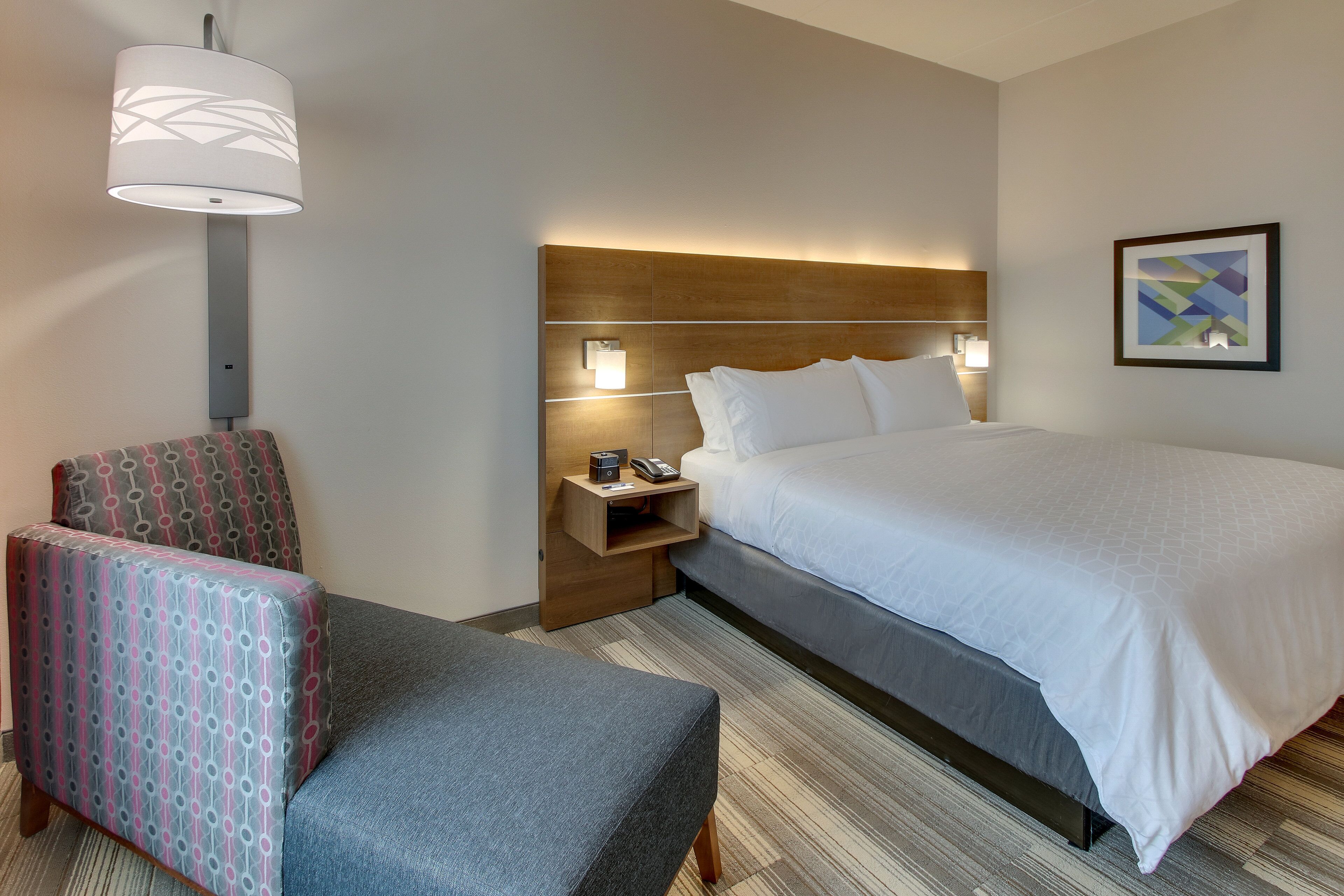 A hotel room with a bed, a chair, a nightstand with a lamp, and a wall art piece.