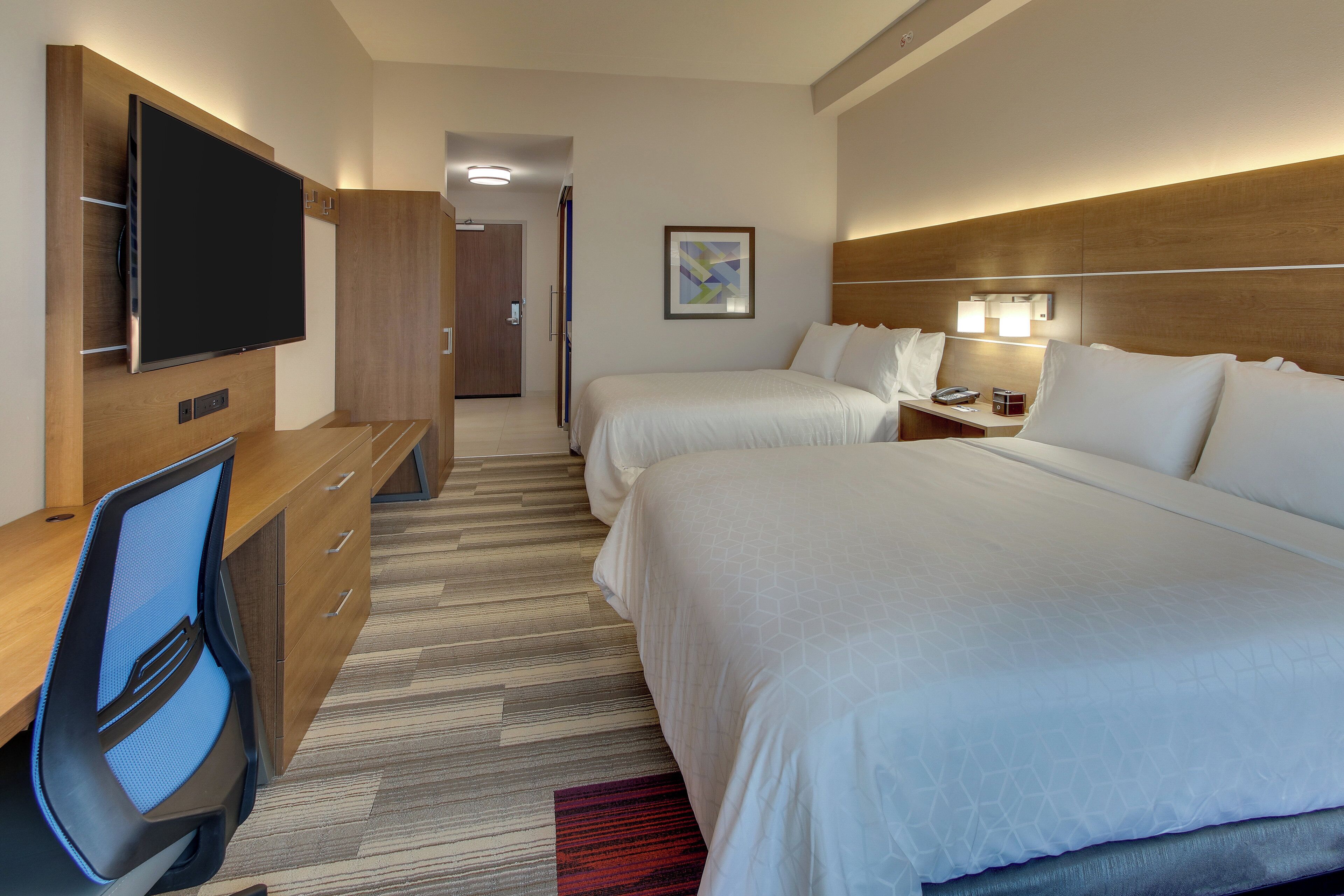 Habitación de hotel con televisión, dos camas, escritorio con silla, mesita de noche con lámpara y puerta que da a otra habitación.