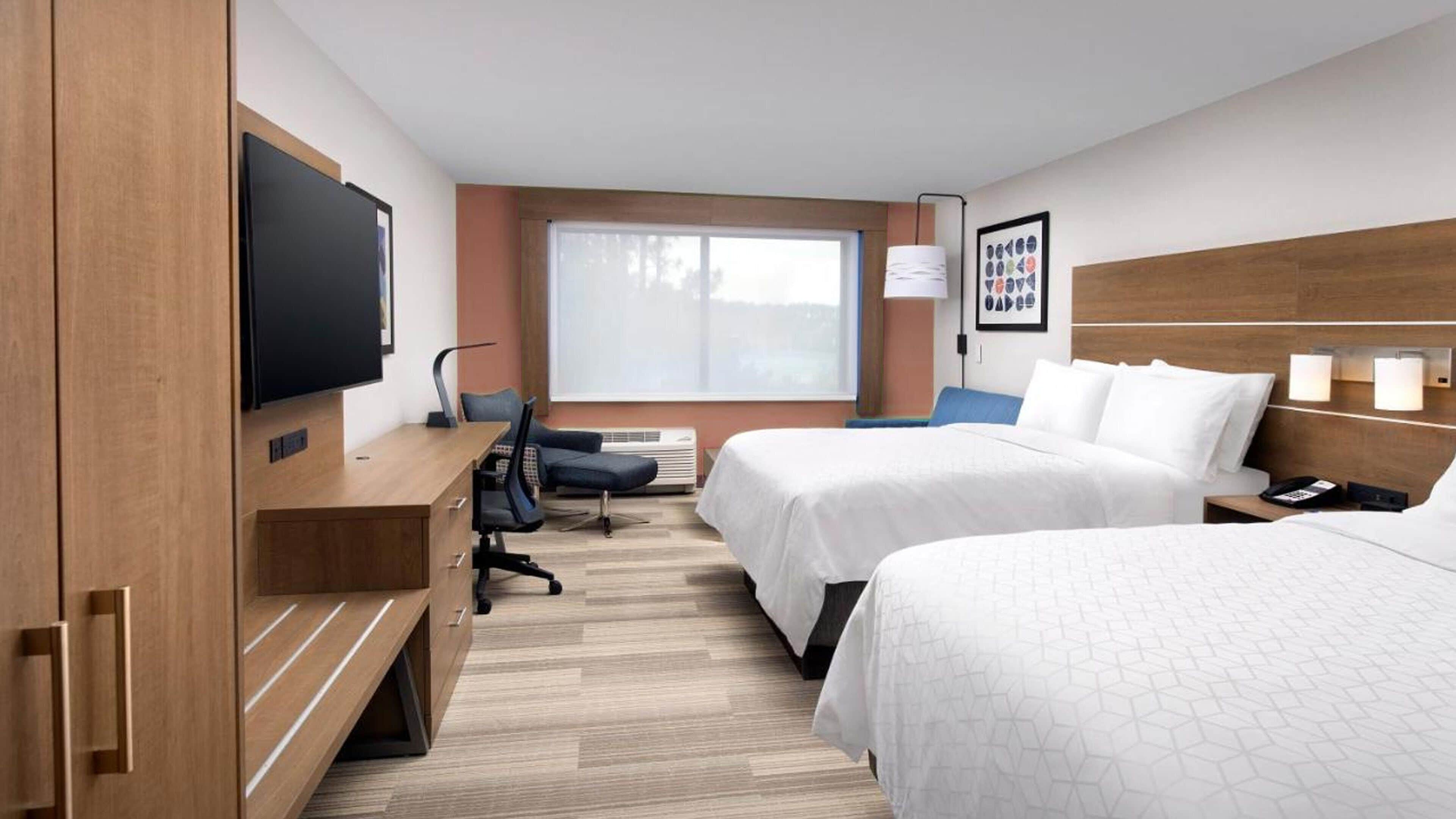 Ein Hotelzimmer mit zwei Betten, einem Schreibtisch mit Fernseher, einem Sessel, einem Fenster und einer Wandlampe.