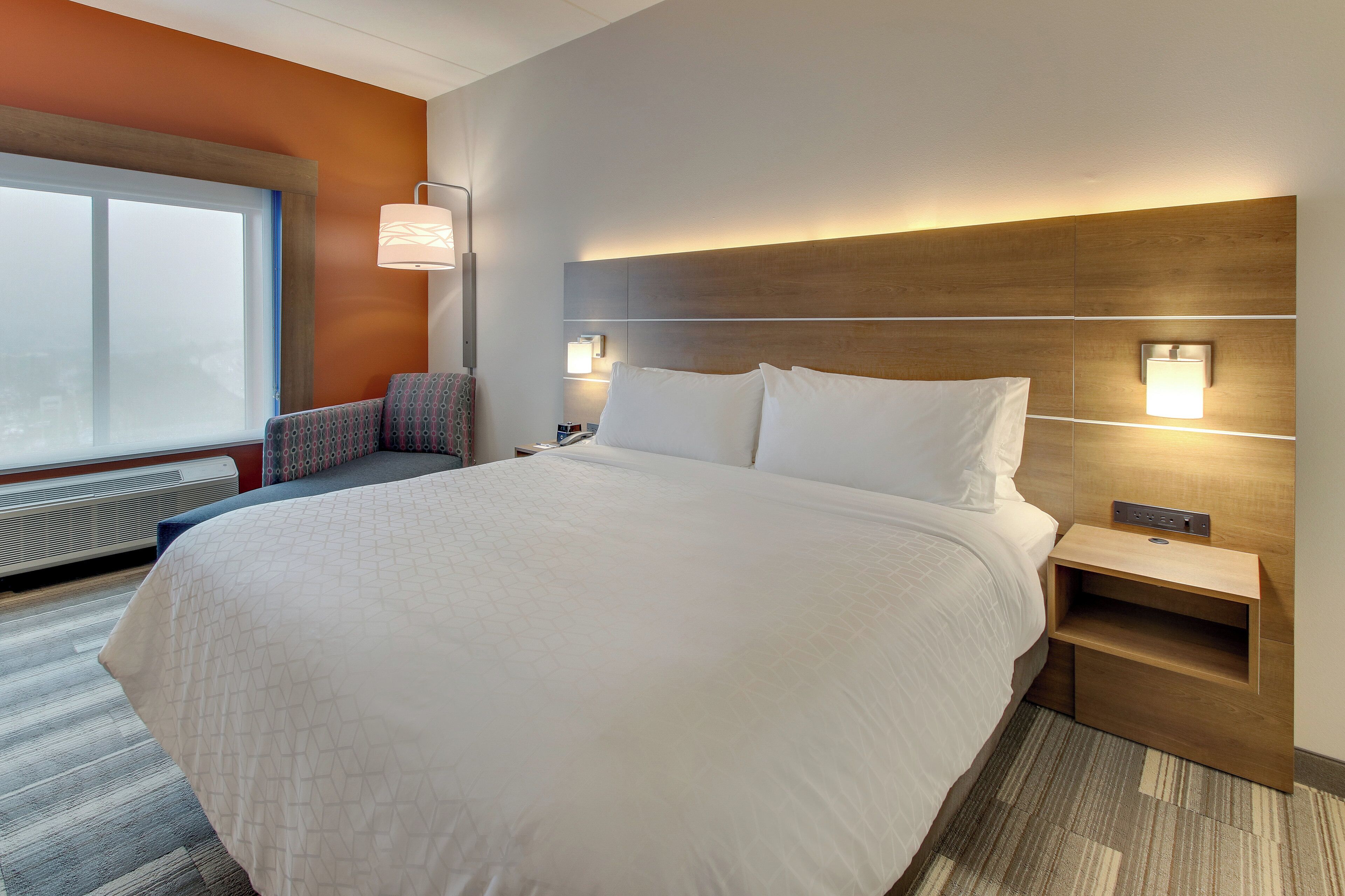 A hotel room with a large bed, a nightstand, a chair, and a window with a view.