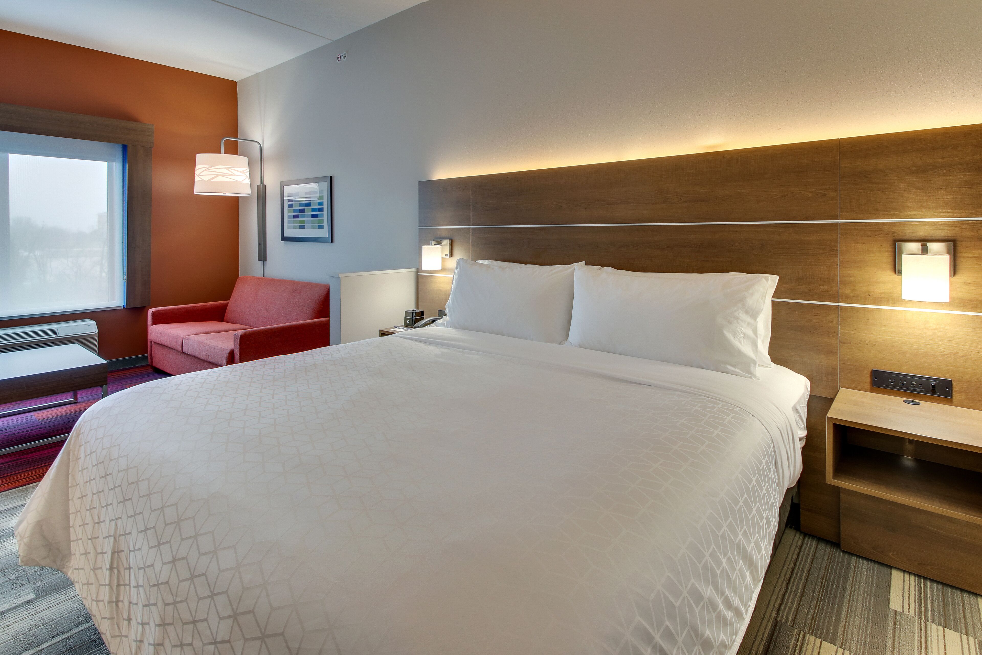 Habitación de hotel con una cama grande, un sillón rojo, una mesita de noche y una lámpara de pared.