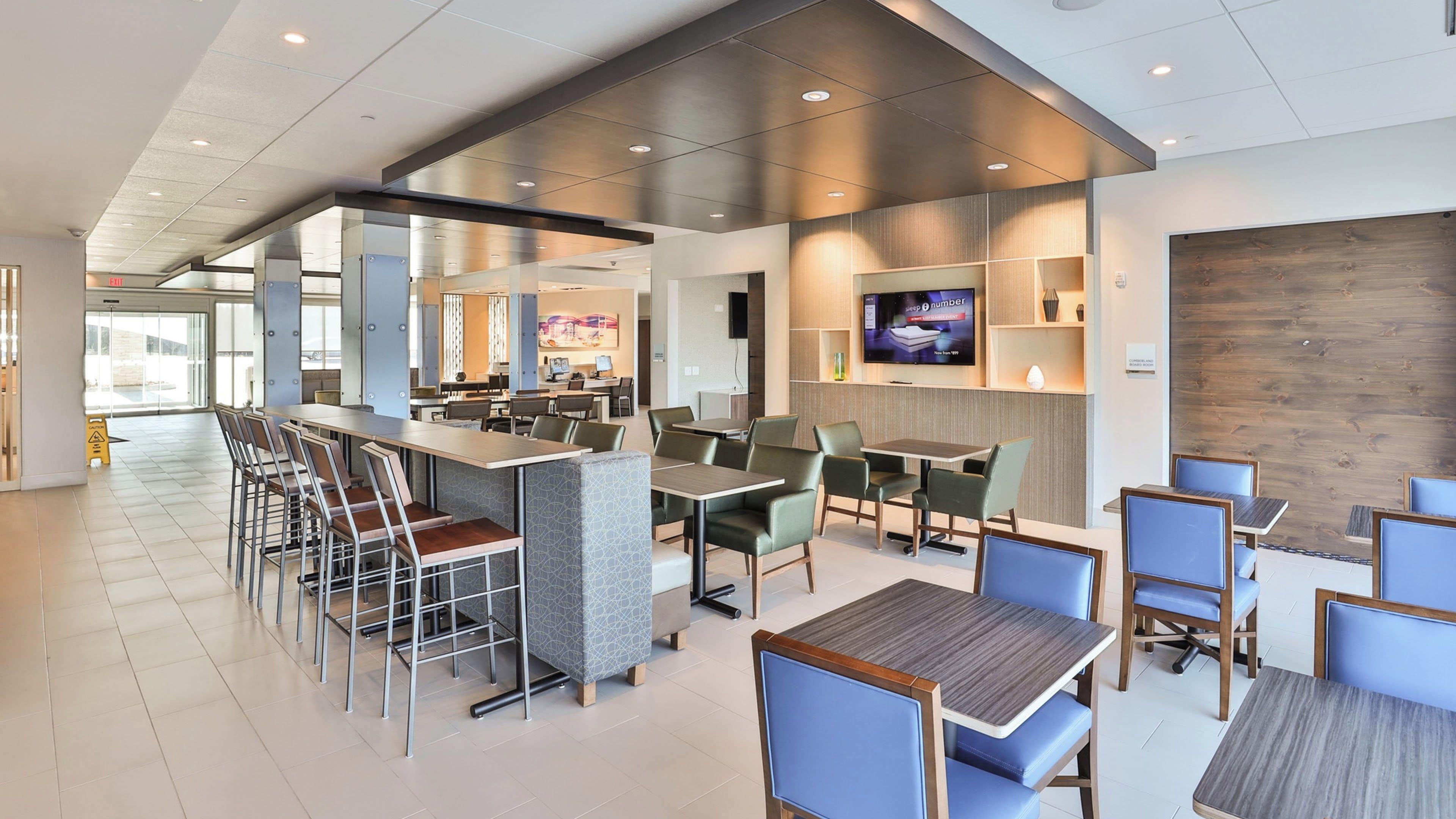 Uma área de refeições moderna, com banquetas de bar e mesas individuais, uma tela de televisão e um painel de madeira decorativo.