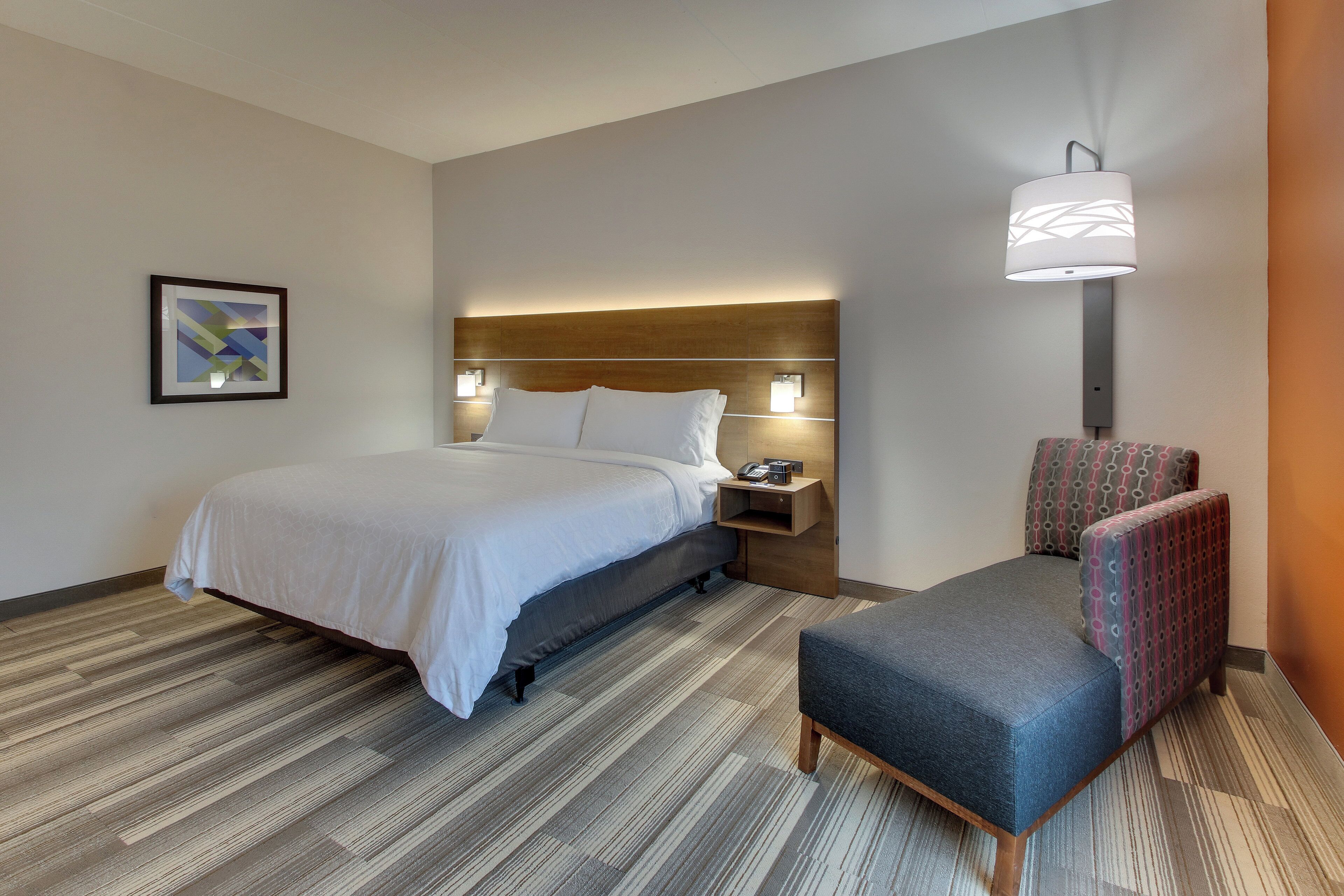 A hotel room with a bed, bedside table, lamp, chair, and a framed picture on the wall.