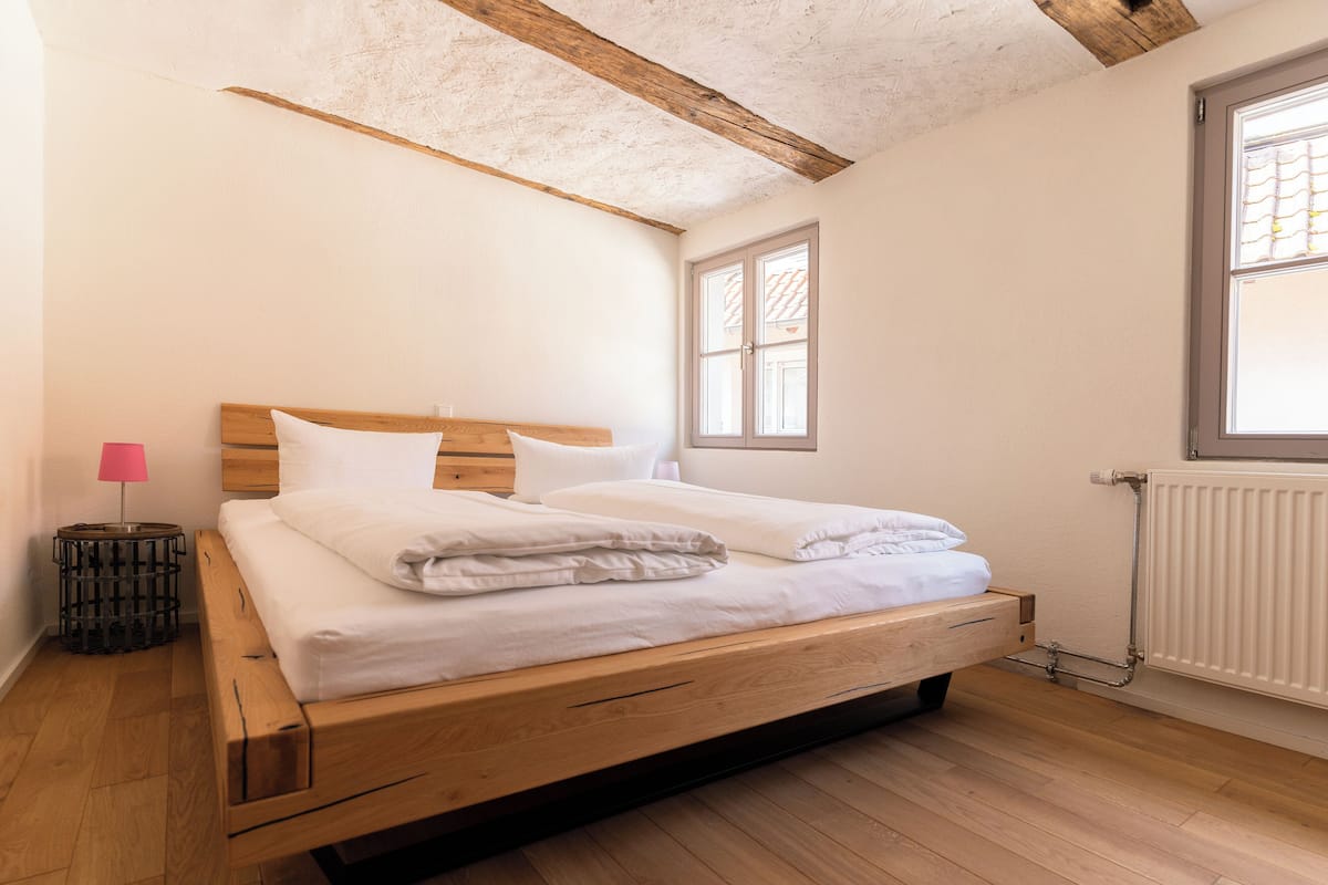 Una cama individual con estructura de madera y ropa de cama blanca, una mesita de noche pequeña con una lámpara rosa y una ventana con persianas.