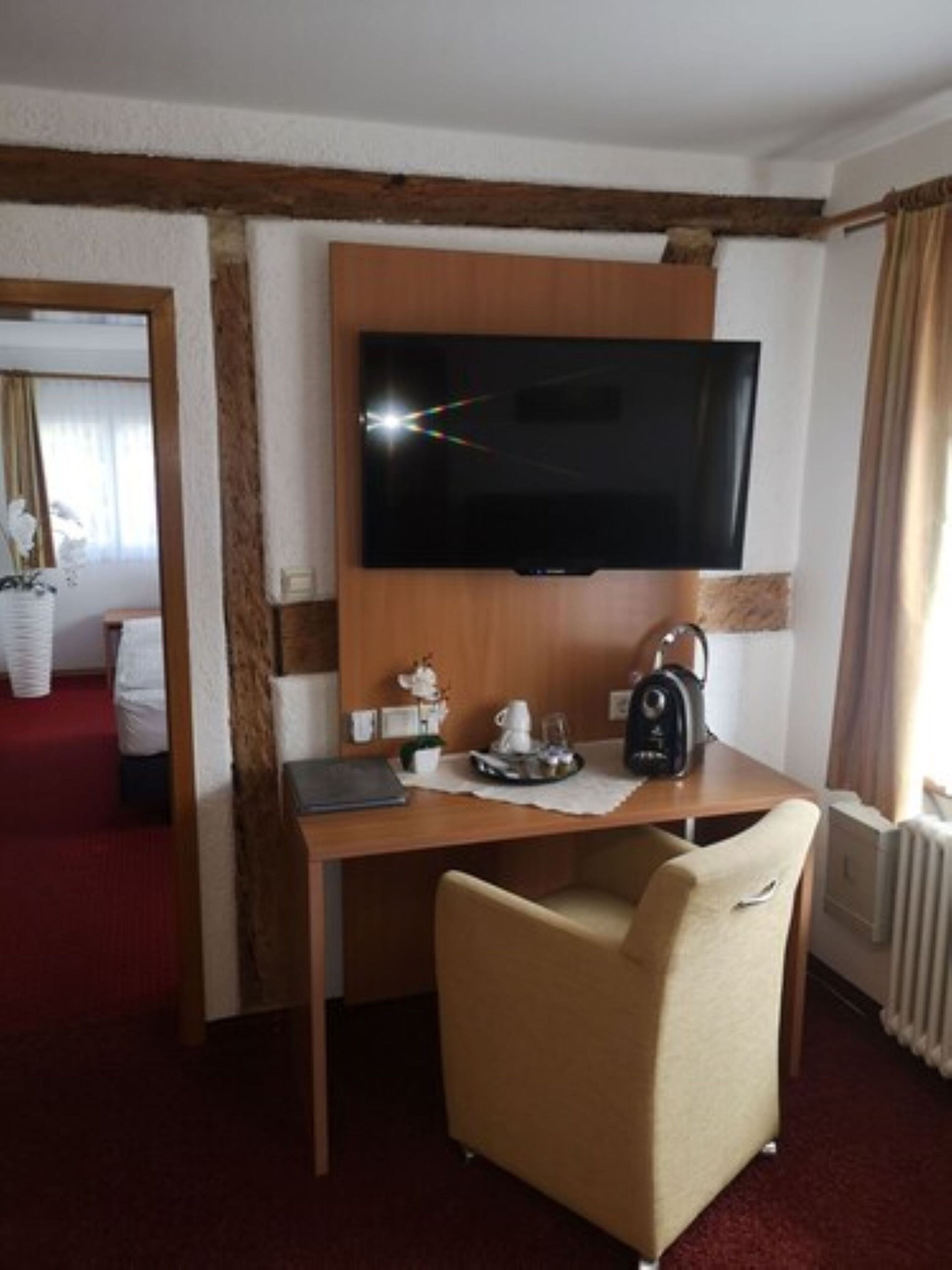 A hotel room with a wooden desk, a flat-screen TV mounted on the wall, a chair, and a small table with a teapot and cups.