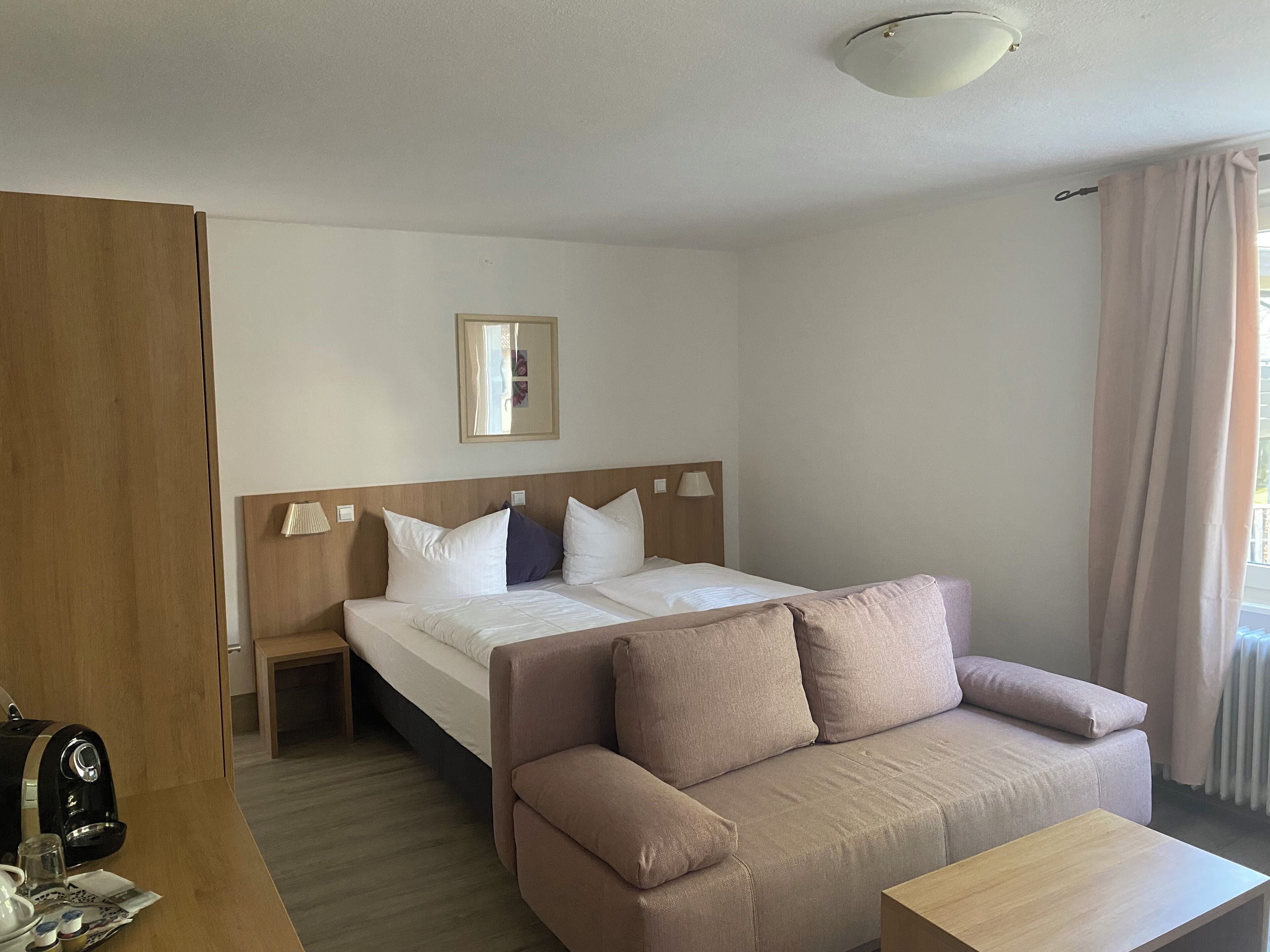 A hotel room with a bed, a wooden headboard, a bedside table with a lamp, a sofa, and a coffee maker.