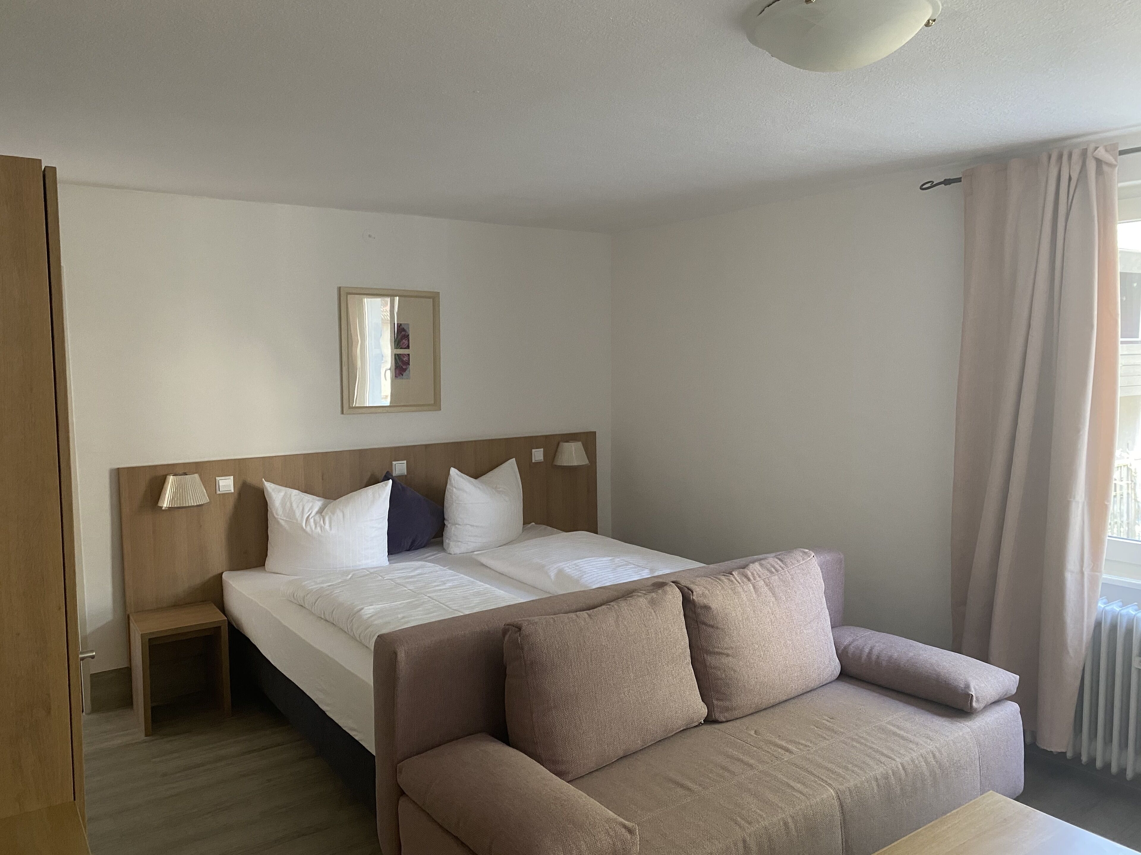 A hotel room with a bed, a wooden headboard, a bedside table, a small nightstand, a sofa, and a window with curtains.