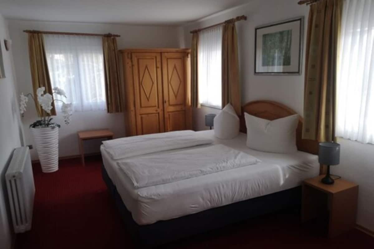 Habitación de hotel con cama doble, armario de madera, mesita de noche con lámpara y planta en maceta.