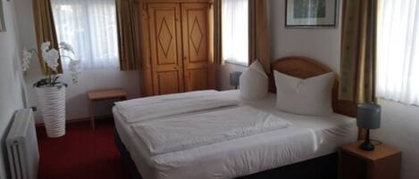 A hotel room with a double bed, a wooden wardrobe, a bedside table with a lamp, and a potted plant.
