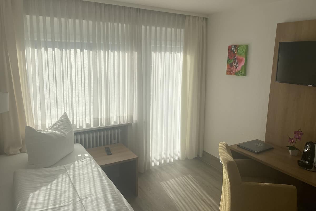Habitación de hotel con cama, escritorio con una computadora portátil, silla, televisor y ventana con cortinas translúcidas.