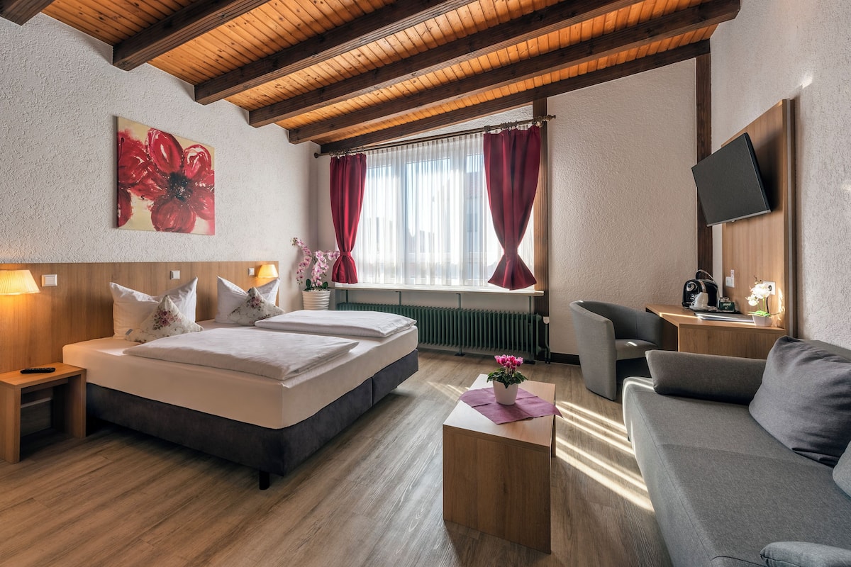 Una habitación de hotel con techo de madera, una cama grande con dos almohadas, una mesita de noche con lámpara, un sofá, un televisor y una ventana con cortinas.