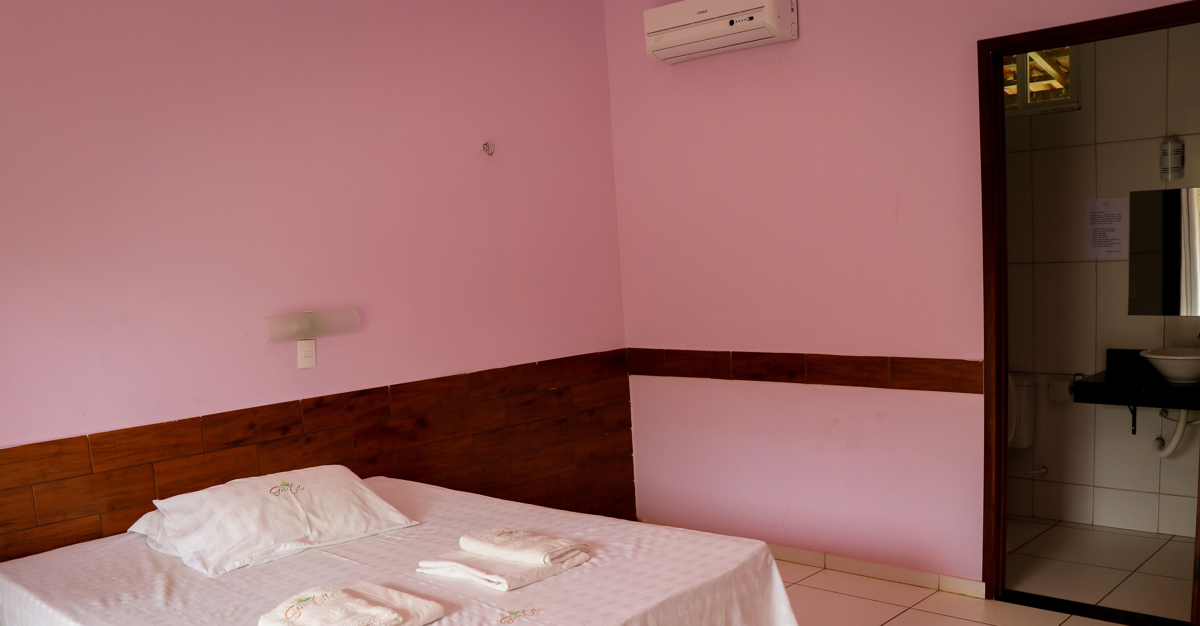 A bedroom with a bed, pillows, a wooden headboard, a wall-mounted air conditioner, and a bathroom visible through an open door.