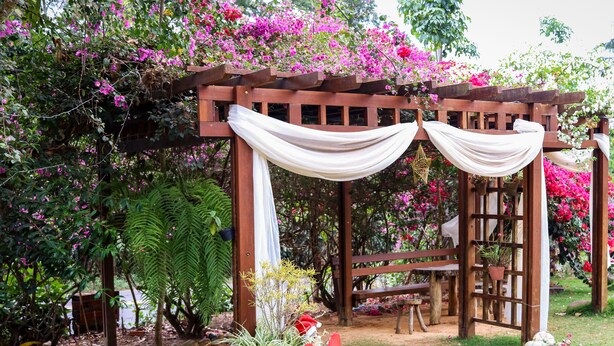 Une pergola en bois, ornée de rideaux blancs, entourée d’une végétation luxuriante et de fleurs roses éclatantes.