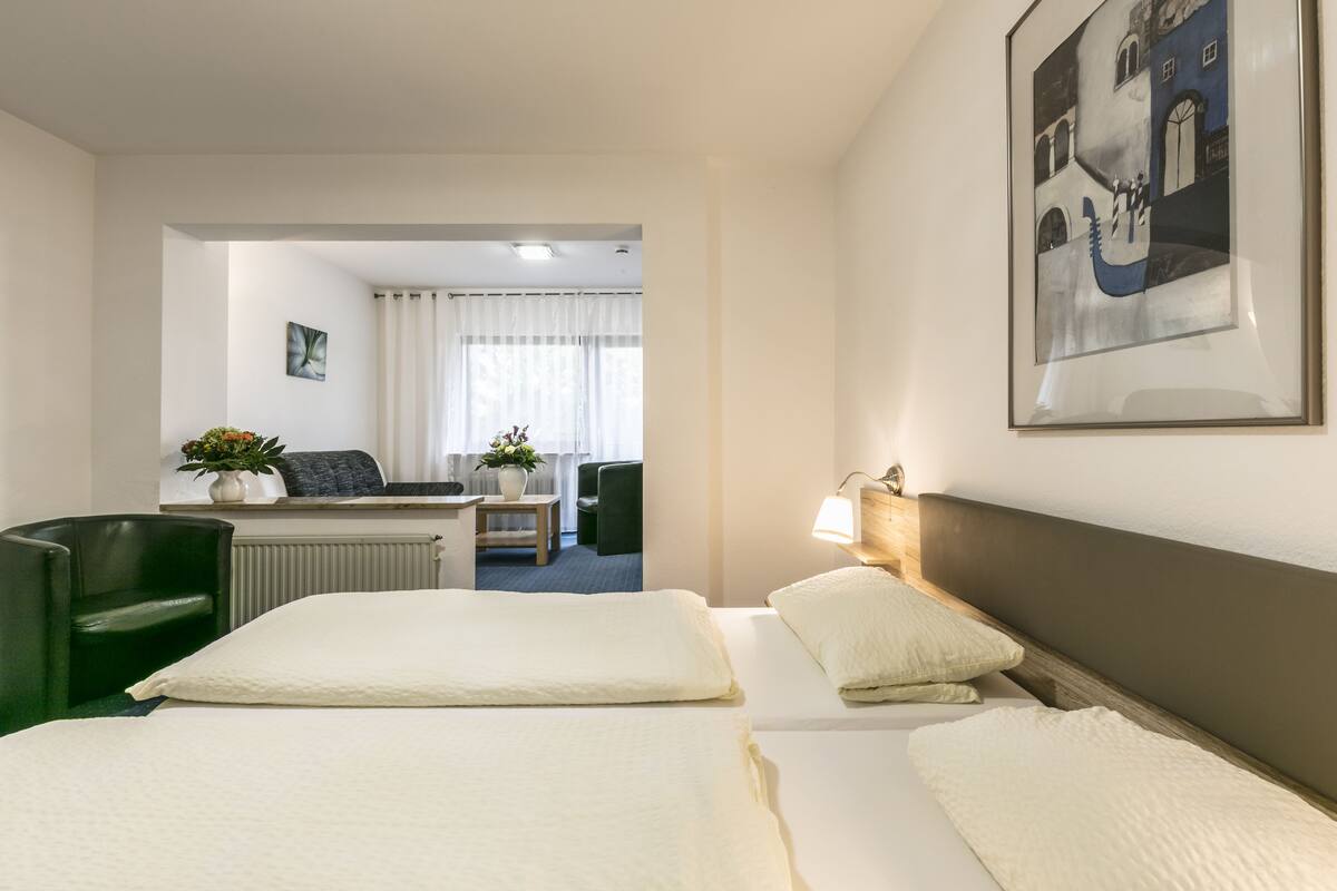 Ein Hotelzimmer mit zwei Betten, einem Schreibtisch, einem Sessel, einer Zimmerpflanze und einem gerahmten Bild an der Wand.