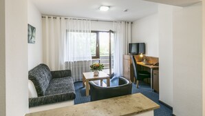 A hotel room with a sofa, a small table with a vase, a desk with a chair, a television, and a balcony with curtains.