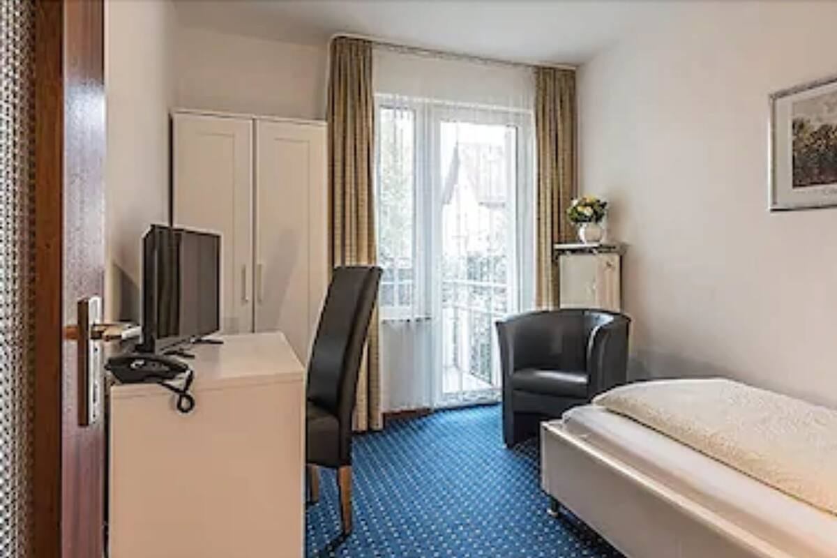 Ein Hotelzimmer mit einem Bett, einem Schreibtisch mit Computer, einem Stuhl und einem Fenster mit Vorhängen.