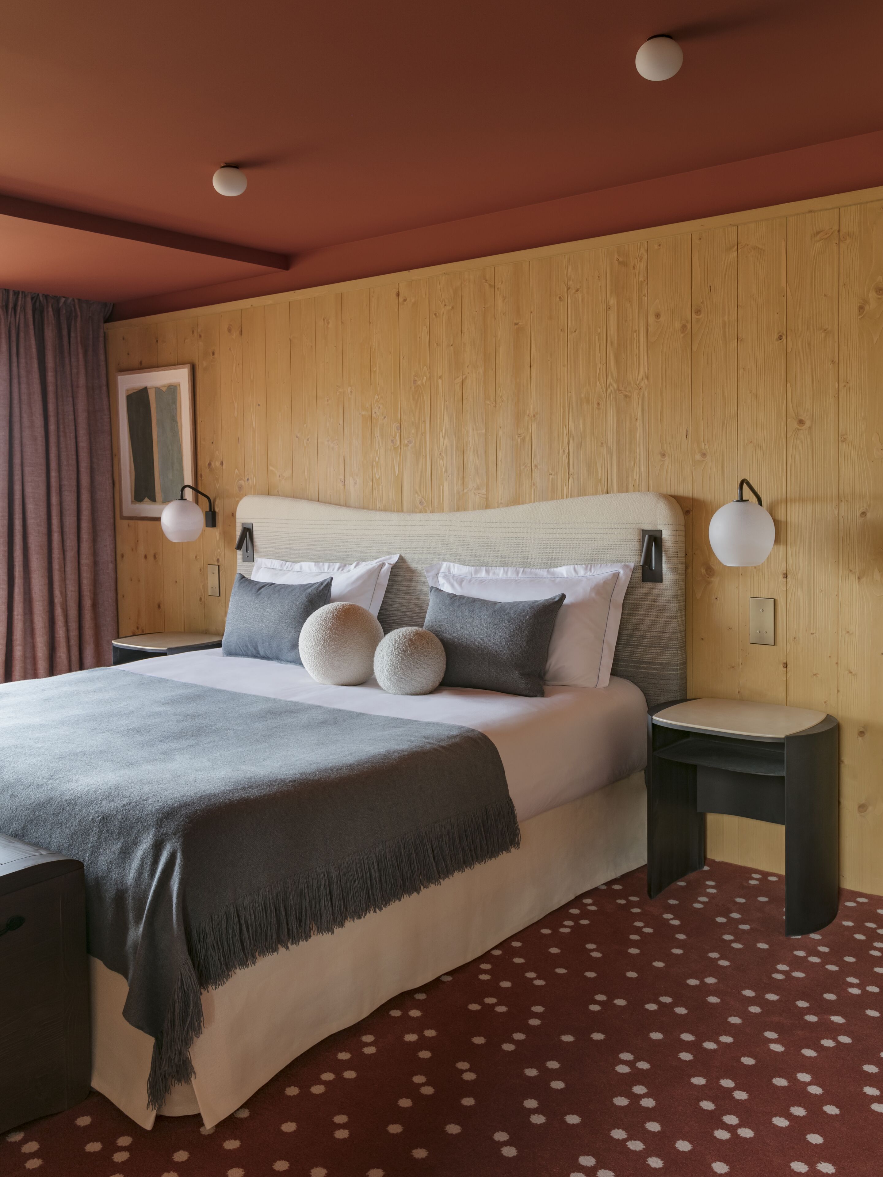 Une chambre à coucher bien aménagée, avec une tête de lit en bois, un lit recouvert d’une couette grise et blanche, deux tables de chevet avec des lampes et un tapis rouge à pois.
