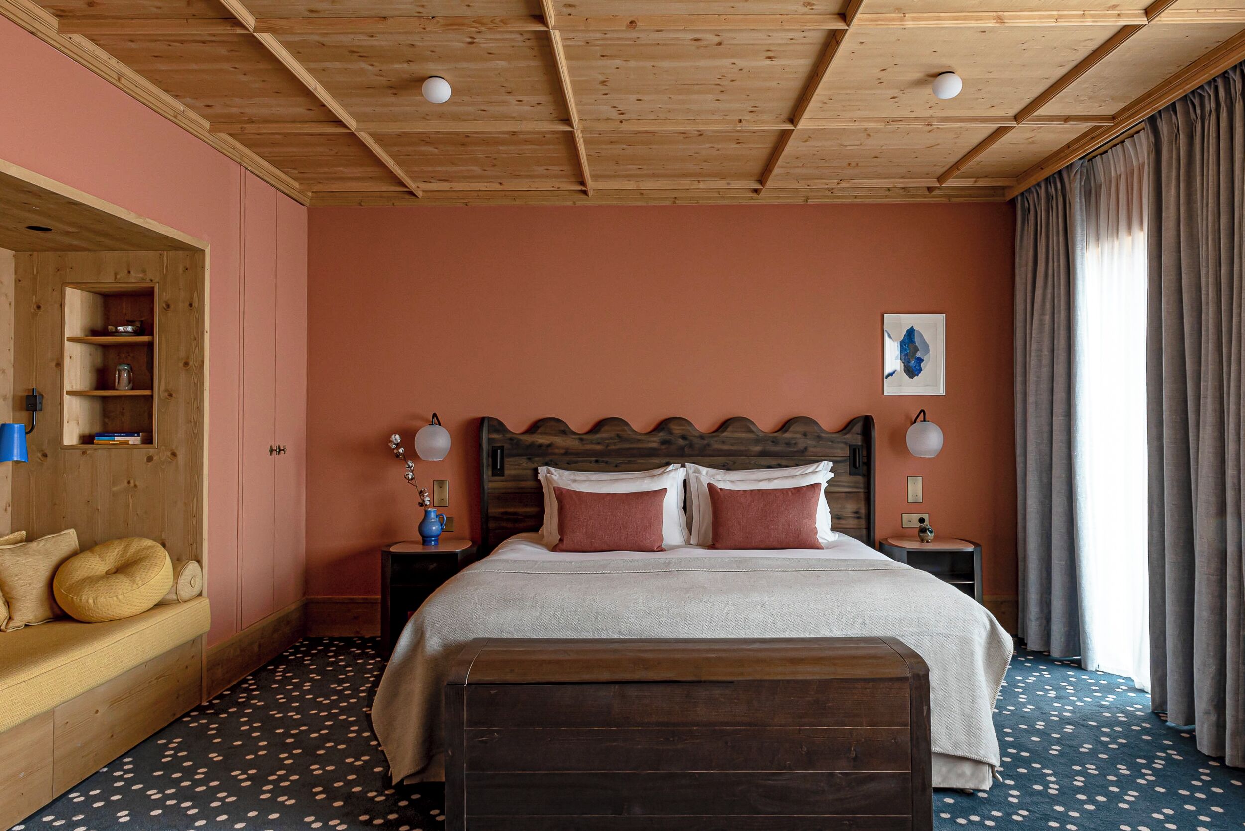 A bedroom with a wooden ceiling, a bed with a wooden headboard, a bench, a nightstand, and a window with curtains.