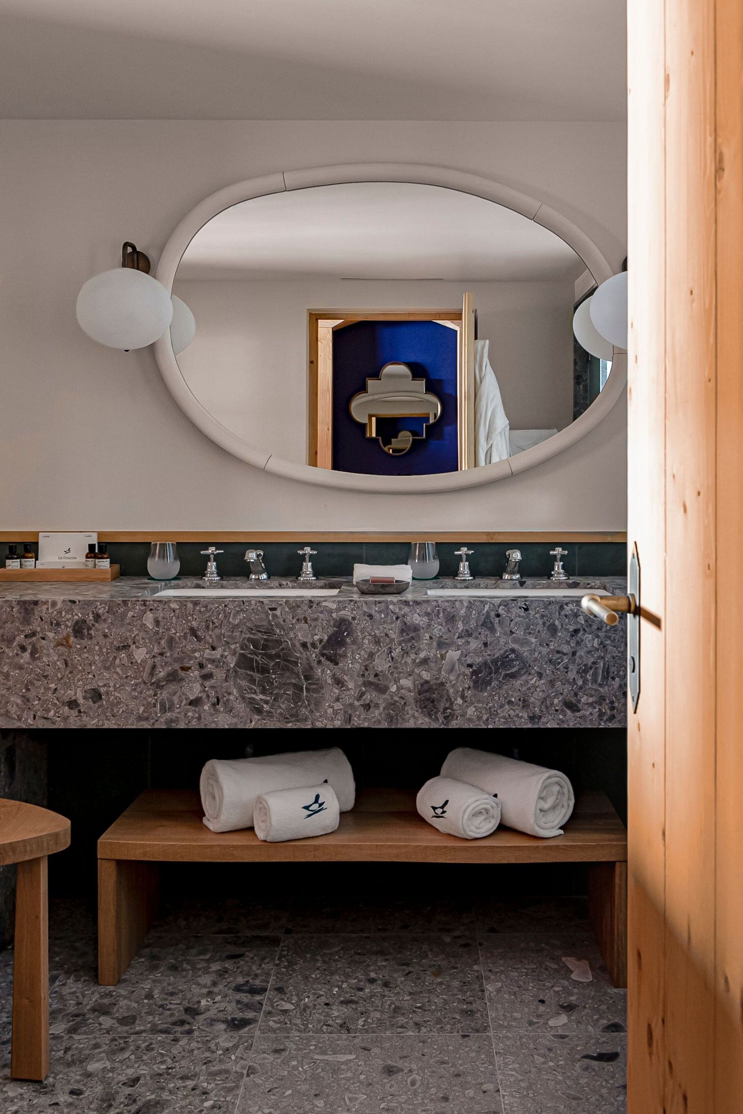 Un baño moderno con lavamanos de mármol, un banco de madera con toallas y un espejo grande con marco redondo.