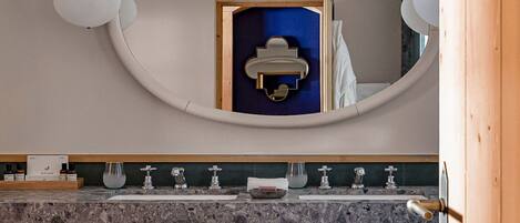 Un baño moderno con lavamanos de mármol, un banco de madera con toallas y un espejo grande con marco redondo.