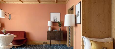 A cozy room with a wooden ceiling, a red sofa, a white chair, a small table with a lamp, and a bed with a yellow pillow and a stuffed toy.