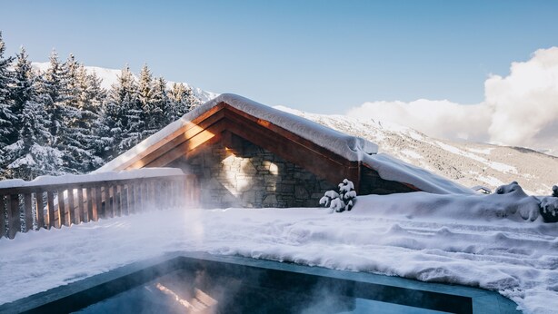 A snow-covered outdoor pool area with a stone building and wooden roof, surrounded by snow-covered trees and mountains.