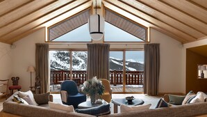 A cozy living room with a high ceiling, wooden beams, a large window with a view of snow-capped mountains, and a balcony with a wooden railing.
