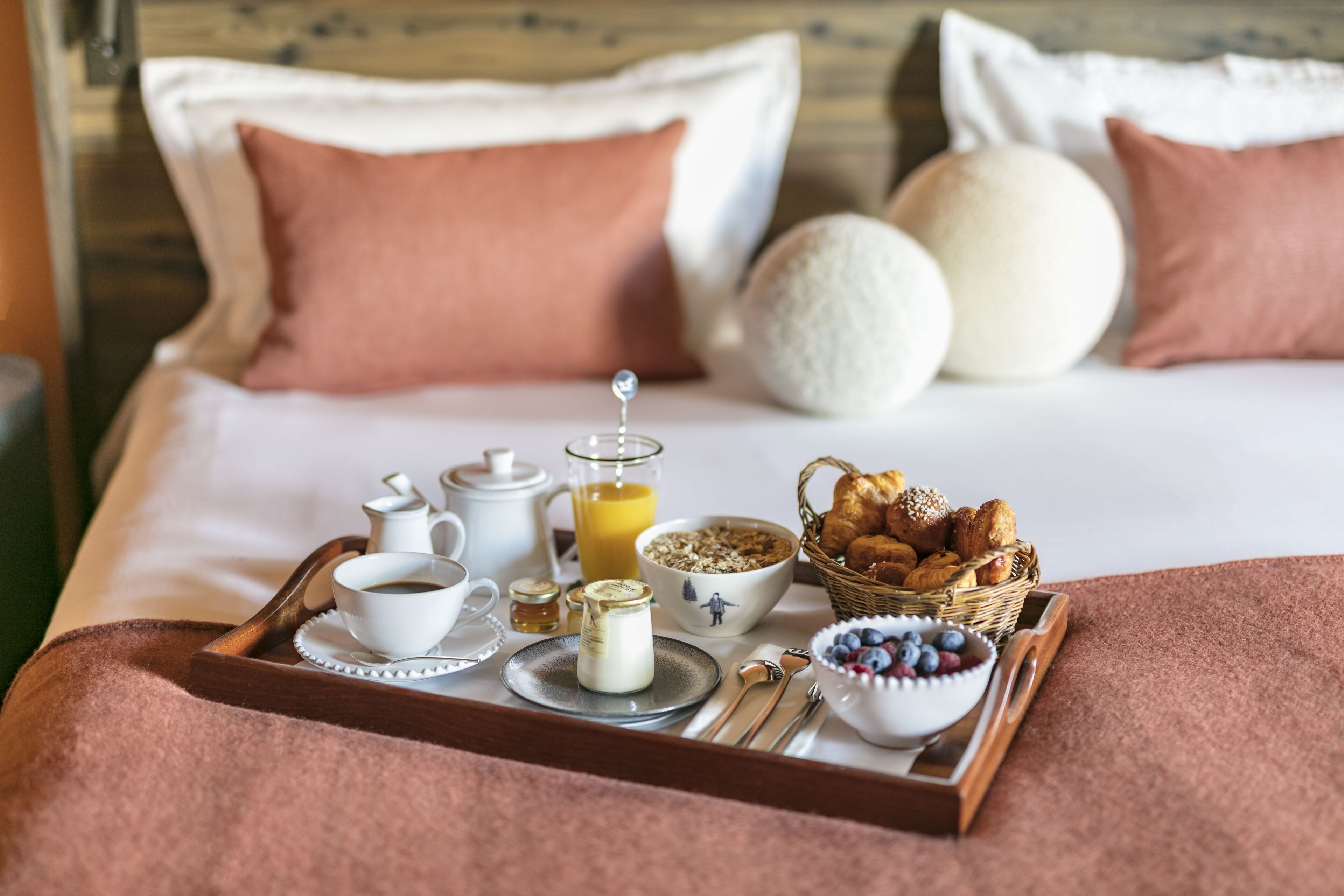En pænt anrettet bakke med morgenmad på sengen, med puder, en kurv med bagværk, en skål med frugt og en kande appelsinjuice.