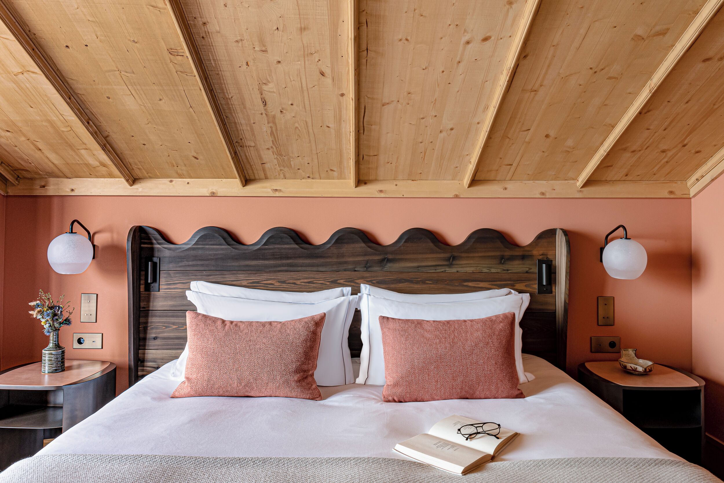 Un dormitorio con techo de madera, cama con ropa de cama blanca y rosa, dos mesitas de noche con lámparas, y un florero con flores sobre una de las mesas.