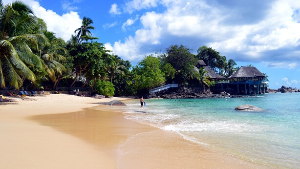 A beach with clear turquoise water, palm trees, and a thatched-roof structure. - Sundown Beach Villa (Mahé Island)