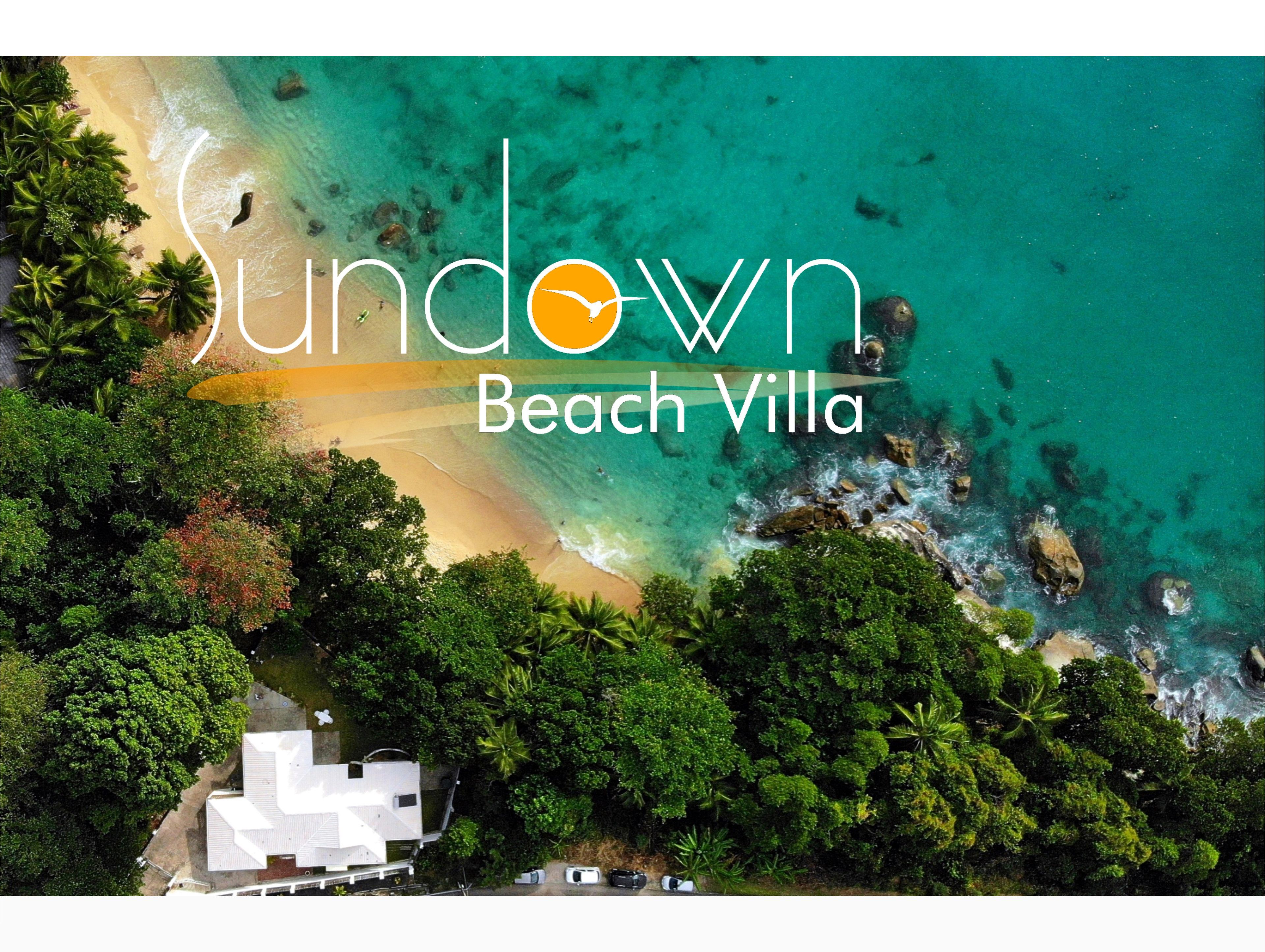 An aerial view of Sundown Beach Villa, featuring a beach with clear turquoise waters, lush greenery, and a white villa structure.