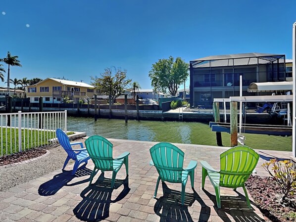 A waterfront patio with green Adirondack chairs, a small table, and a view of a canal with buildings in the background. - 244 Flamingo by Distinctive Beach Rentals (Fort Myers Beach)