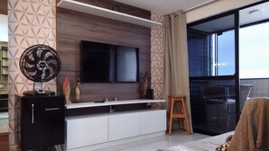 A modern living room with a flat-screen TV mounted on a wooden wall, a black entertainment unit, a standing fan, and a cozy seating area with a blanket. - Apartment with a view of Morro do Careca. (NATAL)
