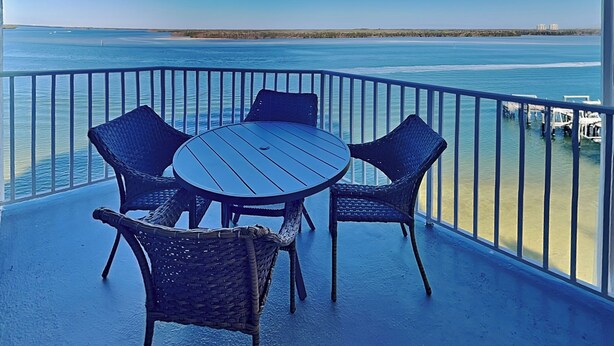 Un balcon avec une table ronde et quatre chaises en osier, donnant sur une plage et l’océan.