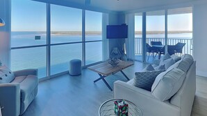 Un vaste salon doté d’une grande fenêtre donnant sur l’eau, d’un ventilateur de plafond, d’un téléviseur à écran plat et d’un coin repas avec des chaises et une table.