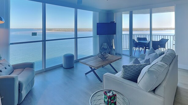 A spacious living room with a large window offering a view of the water, a ceiling fan, a flat-screen TV, and a dining area with chairs and a table. - Lovers Key Resort 404 by Distinctive Beach Rentals (Fort Myers Beach)