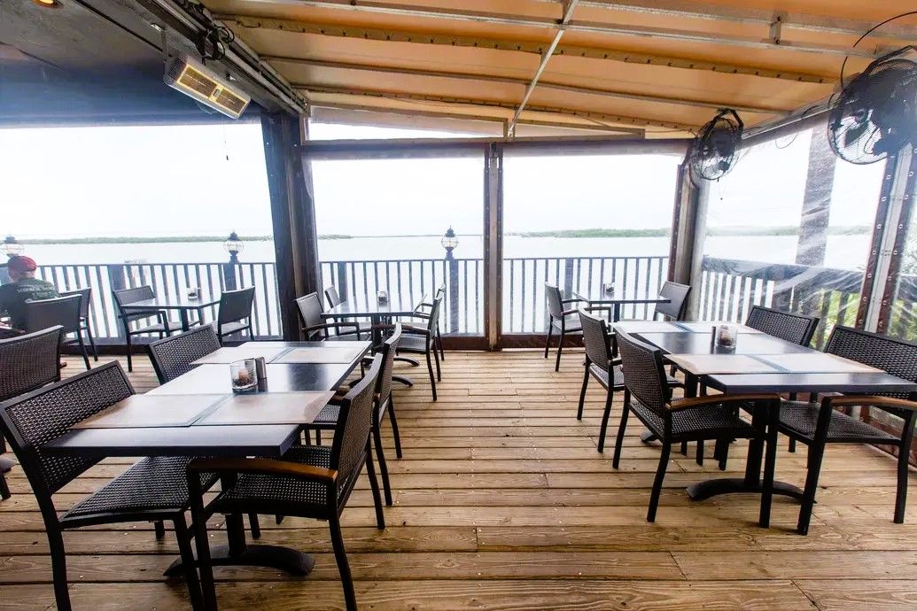 An outdoor dining area with tables and chairs, a wooden deck, and a view of a body of water.